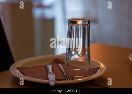 Einrichten. Besteck im Restaurant. Leere Kristallglasreflexion, umgedreht. Messer und Gabel. Runder weißer Teller mit roter Serviette. Stockfoto