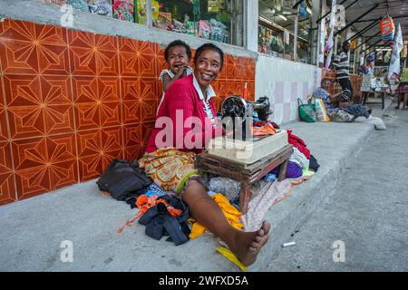 Toliara, Madagaskar – 1. Mai 2019: Unbekannte ältere madagassische Frau, die auf der Straße neben dem Markt sitzt, ein Kind auf dem Rücken weint und Kleidung repariert Stockfoto