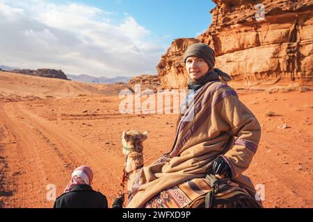 Junge Frau, die in der Wüste Wadi Rum auf einem Kamel reitet, lächelnd über ihre Schulter blickt. Es ist ziemlich kalt, so trägt sie traditionellen Beduinenmantel - bis Stockfoto