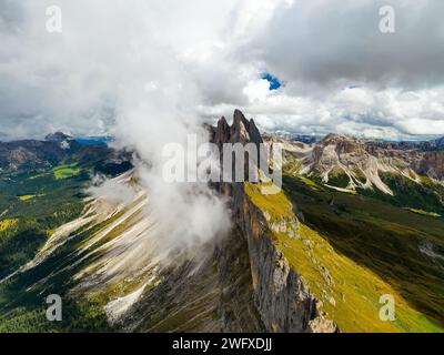 Weiße Wolke umgibt den Riesen Seceda Ridgeline in den italienischen Alpen. Felsige Bergkette mit steilen Hängen unter Cumuluswolken aus der Vogelperspektive Stockfoto