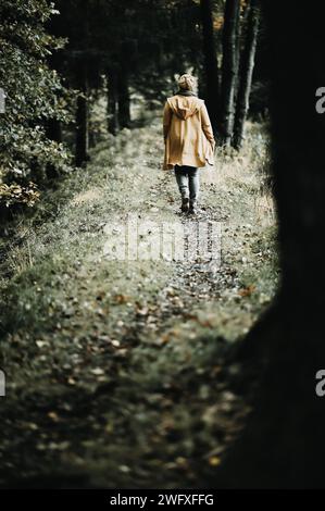 Junge Frau in gelber Strickkleidung in der Natur. Sie läuft auf einem dunklen Waldweg, fotografiert von hinten. Stockfoto