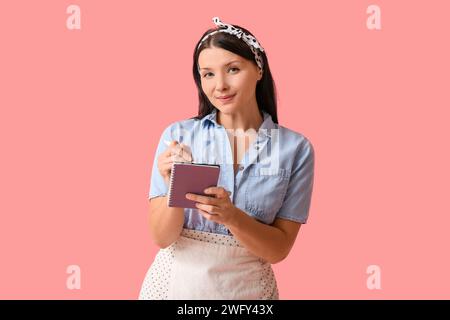 Schöne junge Kellnerin mit Notizbuch auf rosa Hintergrund Stockfoto