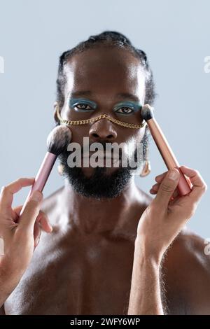 Porträt eines afroamerikanischen schwulen Mannes, der Make-up mit Pinseln aufträgt, die von Crop Artist gehalten werden. Nahaufnahme eines homosexuellen Mannes mit hellem Lidschatten und goldener Kette, der in die Kamera auf blauem Hintergrund blickt. Stockfoto
