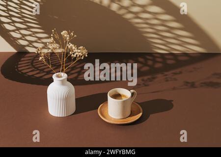 Kreative Komposition aus weißer Vase mit trockenen Blumen und Tasse Kaffee auf braunem und pastellbeigem Hintergrund. Hintergrundbild mit Lichtern und Schatten. Stockfoto