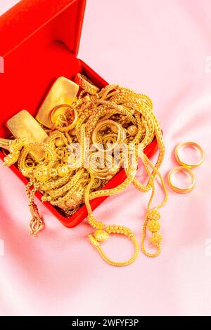 Viele goldene Halsketten und Goldbarren in roter Box auf rosa Samtstoff Hintergrund. Stockfoto