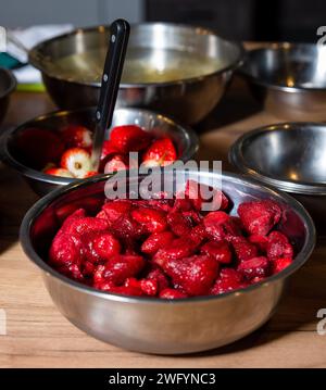 Eine Edelstahlschale voller leuchtend roter Erdbeeren, mit Küchenschalen und einem Spachtel im Hintergrund. Stockfoto