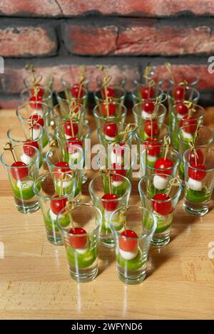 Eine Auswahl an Mini-Caprese-Salat-Vorspeisen mit Kirschtomaten, Mozzarella und Pesto in Shot-Gläsern, die auf einem Holztisch vor einem bri ausgestellt werden Stockfoto