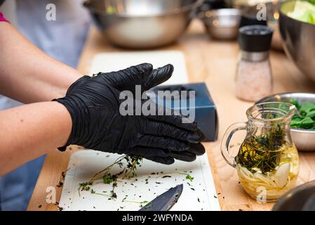 Nahaufnahme der Hände in schwarzen Handschuhen, die einen frischen Kräuteraufguss mit Olivenöl vorbereiten und einen kulinarischen Prozess auf einer Holzoberfläche zeigen. Stockfoto