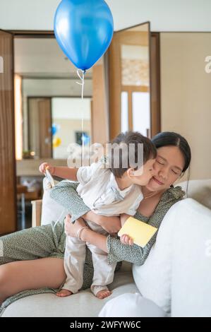 Niedlicher, einjähriger, multirassischer Junge, der seine Mutter küsst, während er seinen ersten Geburtstag mit seiner Mutter feiert und zu Hause einen blauen Ballon trägt Stockfoto