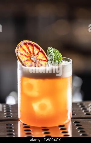 Ein Glas Jungle Bird Cocktail garniert mit getrockneten roten Orangen- und Minzblättern an der Bar. Stockfoto