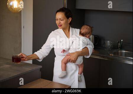 Wunderschöne, ethnische junge Frau in weißem Bademantel, eine liebevolle Happy, die ihr Baby trägt und morgens Tee trinkt, um zum Frühstück zu essen. Personen Stockfoto