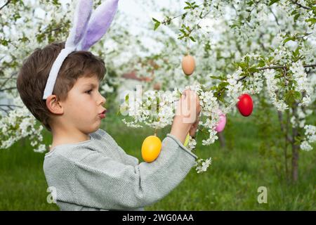 Ostereierjagd. Vorschuljunge, der Hasenohren trägt und bunte Eier auf der Ostereierjagd im Garten findet. Kind, das Zweige eines blühenden Baumes mit verziert Stockfoto