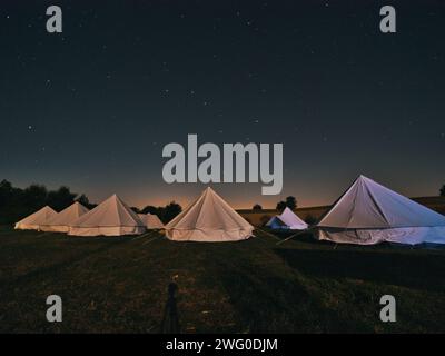 Kreisförmige Anordnung weißer Zelte in einem sternenbeleuchteten Feld, umgeben von einem dunklen Himmel, schaffen eine friedliche und ruhige Atmosphäre unter den hellen Sternen. Stockfoto