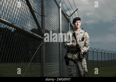 Ein kaukasischer, männlicher Luftwaffe-Sicherheitsmann in Uniform-Posen mit seinem M-4-Gewehr in der Nähe einer gesicherten Zaunlinie mit Stacheldraht. Stockfoto