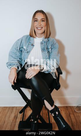 Das kurze blonde Mädchen in jeansjacke sitzt und wartet an der weißen Wand Stockfoto