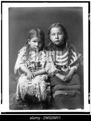 Bull Shoe's Children, 1910. Zwei Mädchen, durchgehend, sitzend, nach vorne gerichtet, mit lockeren Haaren, eines trägt ein Kleid aus Bucklederleder, das mit Elchzähnen verziert ist, das andere trägt ein Kleid aus perlenbesetztem Stoff. Stockfoto