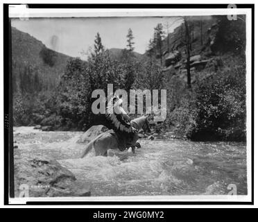 BullChief beim ford, 1905. BullChief trägt eine Kriegshaube und überquert seichte Stromschnellen zu Pferd. Stockfoto