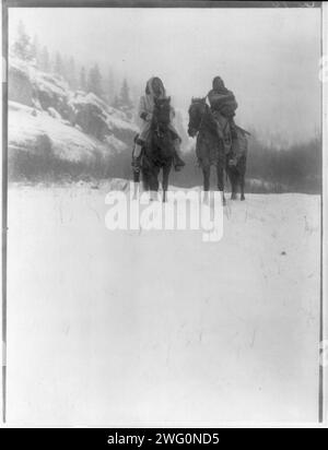 Für eine Winterkampagne - Apsaroke, 1908. Zwei Apsaroke-Männer, einer trägt einen Kapuzenmantel, der andere in eine Decke gewickelt, zu Pferd, auf schneebedecktem Boden, Bäume und Hügel im Hintergrund. Stockfoto