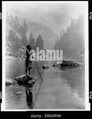 Ein rauchiger Tag im Sugar Bowl-Hupa, 1923. Hupa Mann mit Speer, stehend auf Felsen in der Mitte des Baches, im Hintergrund, Nebel verdeckt teilweise Bäume an Berghängen. Stockfoto