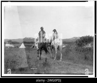 Zwei Töchter eines Häuptlings zu Pferd, die von der Kamera weg zu Zelten im Hintergrund reiten, um 1907. Stockfoto