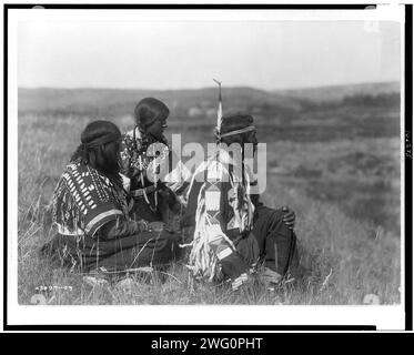 Mit Blick auf das Camp-Piegan, 1910. Ein Mann und zwei Frauen sitzen auf einem Hügel, nach rechts gerichtet. Stockfoto