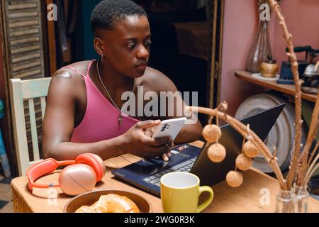 Junge kurzhaarige Oofro-Frau aus der Ahitianischen Ethnie, die zu Hause sitzt und online mit ihrem Handy arbeitet und Laptop benutzt, mit einer Tasse Kaffee und Croissants Stockfoto