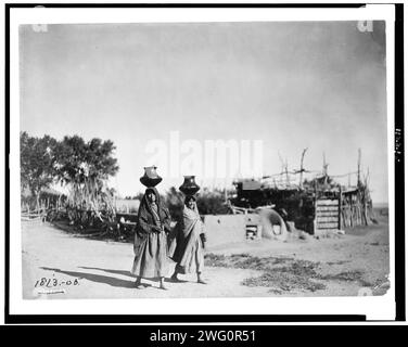 Im Dorf Santa Clara, 1905. Zwei Pueblo-Frauen, die Krüge auf dem Kopf tragen; Bäume, Zugzaun, Stockwerk entlang des unbefestigten Weges im Hintergrund. Stockfoto