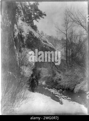 Vorbei an der Klippe Apsaroke, um 1908. Zwei Apsaroke zu Pferd machen sich auf den Weg entlang eines flachen Baches neben einer Klippe. Stockfoto