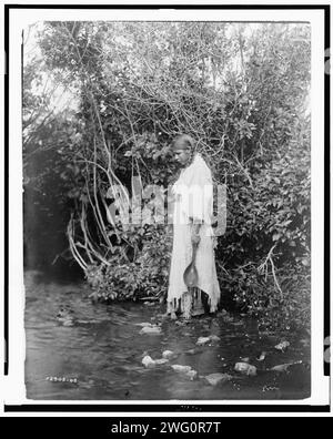 Am Ufer Arikara, 1908. Junger Arikara-Indianer, der im flachen Wasser steht, in einem Kleid aus Buckelleder, mit Bäumen im Hintergrund, North Dakota. Stockfoto