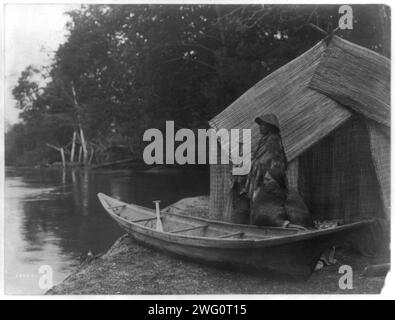 Angellager Skokomish, 1913. Skokomish Paar vor dem Haus aus Schilfmatten, einer sitzt neben dem Kanu, das auf das Ufer gezogen wurde, und der andere steht mit einem Paddel. Stockfoto