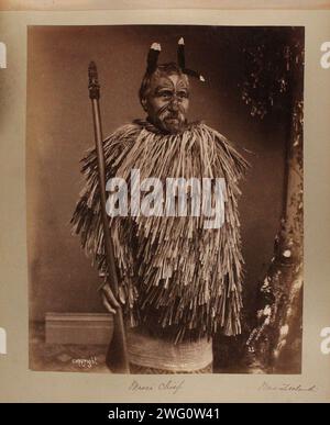 Maori-Häuptling Menehira posiert stehend. Er hat einen Schnurrbart, kurzes, graues Haar mit Federschmuck und ausgedehnten Gesichtstattoos. Er trägt einen Mantel aus Lagen von Flachsfransen um seinen Oberkörper und hält einen Holzstab mit einer Schnitzerei oben. Porträt mit Dreiviertellänge. Von Charles Appleton Longfellow's Reise nach Neuseeland im Jahr 1891. Stockfoto