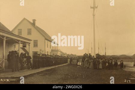 Matrosen stehen an der Promenade mit einer Gruppe einheimischer Frauen und Männer in der Nähe, die die Zeremonie beobachten, möglicherweise Iliulkuk (Unalaska) auf Unalaska Island, Alaska/, 1894 oder 1895. In Album: Dokumentarfotos von Alaska und den Inseln vor der Nordküste, 1894-1895, l. 68, Nr. 317. Stockfoto