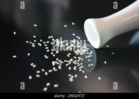 Wohnmöbel, Stil und Essen, weiße Keramikvase mit weißen Sesamsamen auf schwarzem glänzenden Hintergrund. Stockfoto