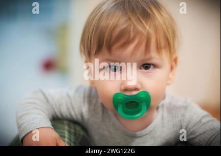 Der süße Junge blickt direkt in die Kamera, die Neugier in seinen Augen, mit einem leuchtend grünen Schnuller im Mund vor einem sanft fokussierten Innenraum Stockfoto