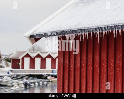 Eiszapfen glitzern, während sie am Rand der traditionellen roten Hütten neben einem ruhigen, schneebedeckten Dock hängen. Stockfoto