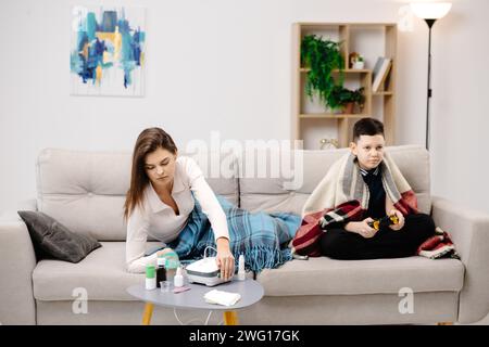 Kranke Dame, die durch Inhalator-Maske zu Hause einatmet, und lustiger Junge mit Gamepad, der Videospiel zu Hause in ihrer Nähe spielt. Stockfoto