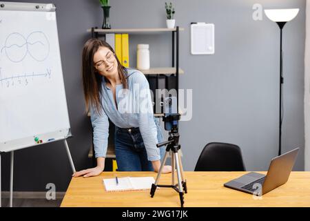 Positive Bloggerin, die auf einem Stativ an der Smartphone-Webcam spricht, einen Beitrag für den Vlog aufnimmt. Online-Lehrer, Coach, Webinar zu Hause. Pressen Stockfoto