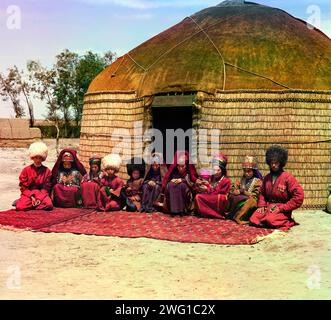 Gruppe von elf Erwachsenen und Kindern, die zwischen 1905 und 1915 auf einem Teppich vor einer Jurte sitzen. Gruppe von Menschen, möglicherweise Turkman oder Kirgiz, in traditioneller Kleidung. Eine Jurte ist ein tragbares Zelt, das von den nomadischen Völkern Zentralasiens zur Unterbringung genutzt wird. Nach der Eroberung Turkestans Mitte der 1800er Jahre übte die russische Regierung starken Druck auf die nomadischen Völker aus, einen sesshaften Lebensstil anzunehmen und sich dauerhaft in Dörfern, Städten und Städten niederzulassen. Stockfoto