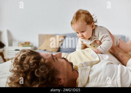 Mann mit lockigen Haaren und Bart, der mit seinem kleinen Sohn auf einem Bett spielt und sich zwischen Vater und Kind verbündet Stockfoto