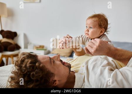 Mann mit lockigen Haaren und Bart, der Hände hält, mit seinem kleinen Sohn mit blauen Augen im Schlafzimmer Stockfoto