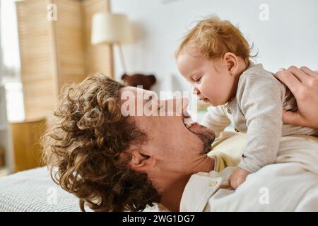 Glücklicher Mann mit lockigen Haaren und Bart, der seinen kleinen Sohn im Schlafzimmer ansieht, Liebe und Pflege des Vaters Stockfoto