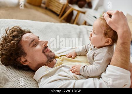 Glücklicher Mann mit lockigen Haaren und Bart, der Haare streichelt, mit seinem kleinen Sohn im Schlafzimmer, Liebe und Pflege Stockfoto