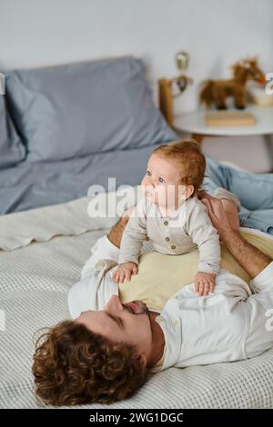 Mann mit lockigen Haaren und Bart, der seinen kleinen Sohn auf einem Bett umarmt und sich zwischen Vater und Kind verbündet Stockfoto