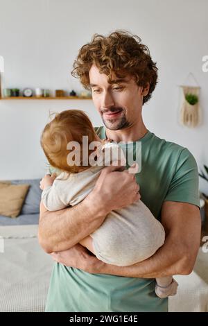 Liebevoller Vater mit lockigen Haaren und Bart, der seinen Jungen in Babykleidung in den Armen hält Stockfoto