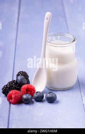 Im Vordergrund, auf dem Holztisch, einige frische Beeren und ein Glas mit reinem und fettarmem Joghurt mit einem Keramiklöffel Stockfoto