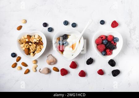 Drei herzförmige Schüsseln auf leicht strukturiertem Hintergrund mit Beeren, getrockneten Früchten, weißem Joghurt, Vollkornflocken und Honig Stockfoto