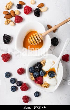 Zwei herzförmige weiße Keramikschalen auf leicht strukturiertem Hintergrund mit Beeren, getrockneten Früchten, weißem Joghurt, Müsli und Honig Stockfoto