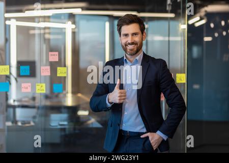 Erfolgreicher und lächelnder junger Geschäftsmann, der in die Kamera blickt und ein Superzeichen mit dem Finger zeigt und die Hand in der Tasche hält. Selbstbewusst in einem Anzug im Büro stehen. Stockfoto