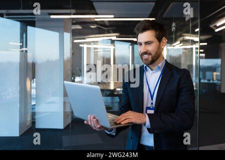 Nahaufnahme eines lächelnden jungen Geschäftsmannes. Ein spezialisierter Programmierer, der bei einer Konferenz in einem Anzug und mit einem Ausweis im Büro steht, einen Laptop hält und benutzt. Stockfoto