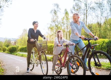 Lächelnder Vater und lächelnde Mutter mit Tochter während der Sommerfahrradrundfahrt. Sie genießen die Zusammengehörigkeit im Sommer-Stadtpark. Glückliche Elternschaft und ch Stockfoto